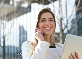 Confident businesswoman using her tablet and phone, smiling outdoors in sunlight.